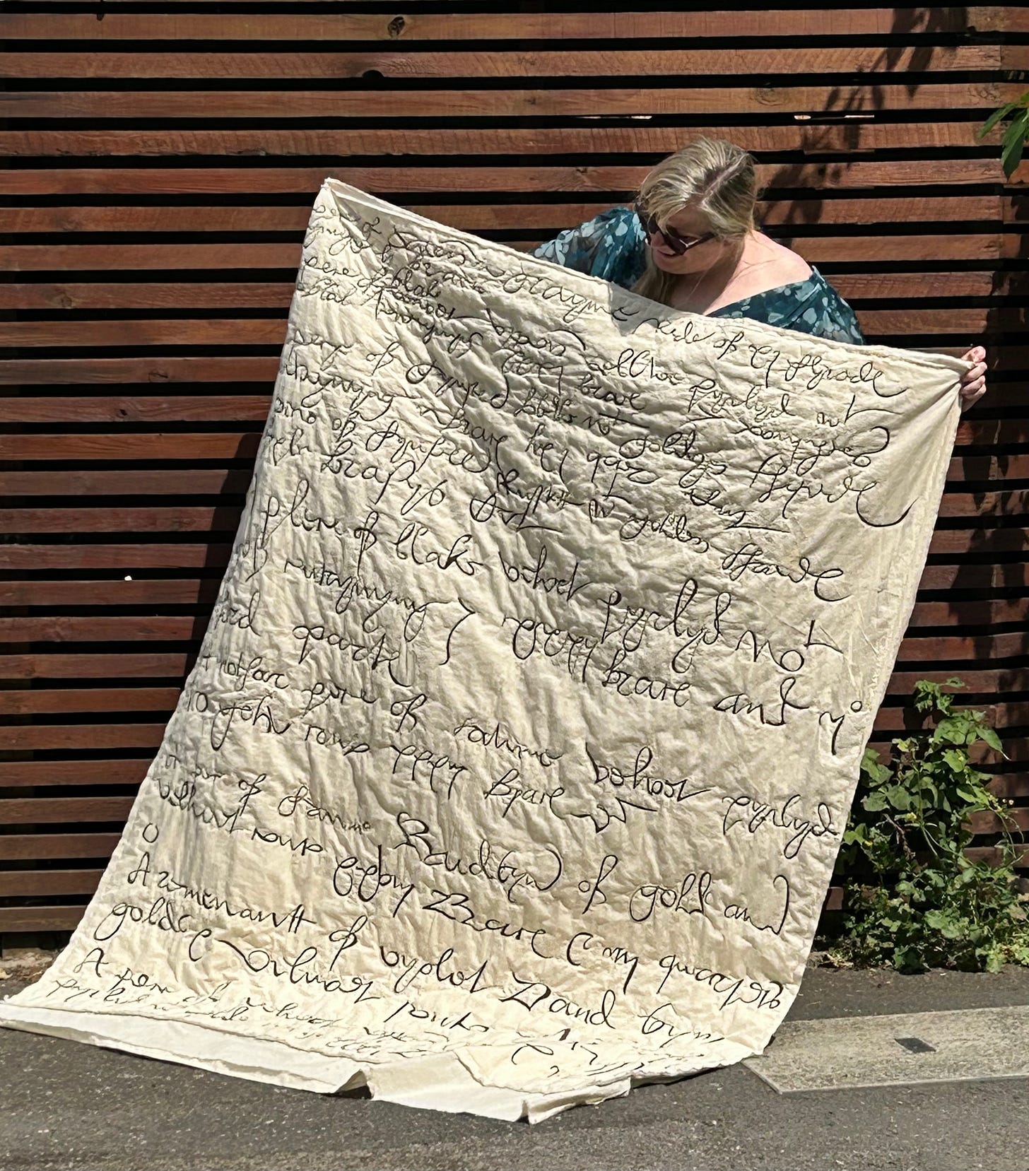 A white woman holds up a large quilted piece decorated with 16th century writing in large scale. 