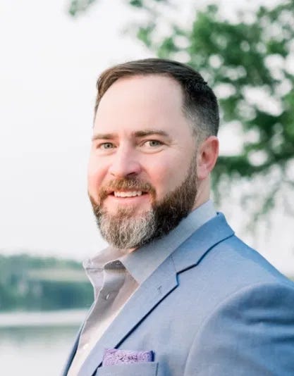 The image shows a man with short brown hair and a full beard, smiling at the camera. He is wearing a light blue suit jacket with a pocket square and a collared shirt. The background features an outdoor setting with water and green trees, creating a serene and natural atmosphere. There are no noticeable landmarks, brands, or recognizable figures in the scene. The image shows a man with short brown hair and a full beard, smiling at the camera. He is wearing a light blue suit jacket with a pocket square and a collared shirt. The background features an outdoor setting with water and green trees, creating a serene and natural atmosphere. There are no noticeable landmarks, brands, or recognizable figures in the scene.