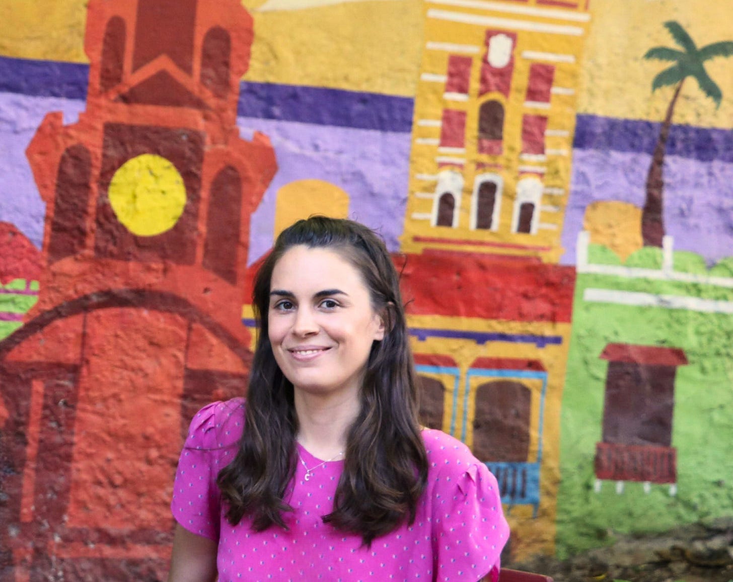Caitlin with long hair wearing a bright pink dress sits in front of a colourful mural depicting buildings