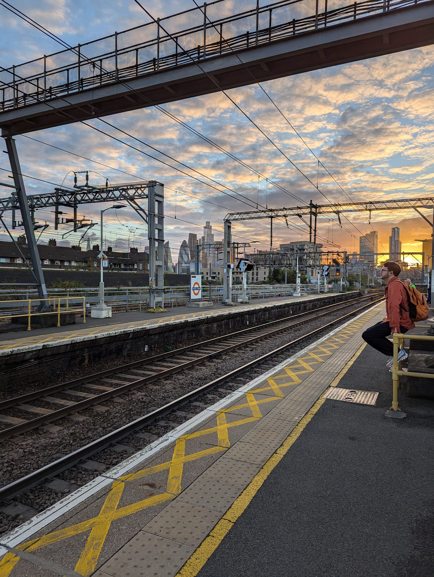 A lone figure in an orange jacket and backpack sits on a yellow railing at an East London Overground platform, watching a dramatic sunset over the City of London skyline — including the Gherkin and other skyscrapers — visible through a web of overhead railway cables and gantries. The sky is streaked gold and orange. Yellow cross markings line the platform edge in the foreground. A lone figure in an orange jacket and backpack sits on a yellow railing at an East London Overground platform, watching a dramatic sunset over the City of London skyline — including the Gherkin and other skyscrapers — visible through a web of overhead railway cables and gantries. The sky is streaked gold and orange. Yellow cross markings line the platform edge in the foreground.