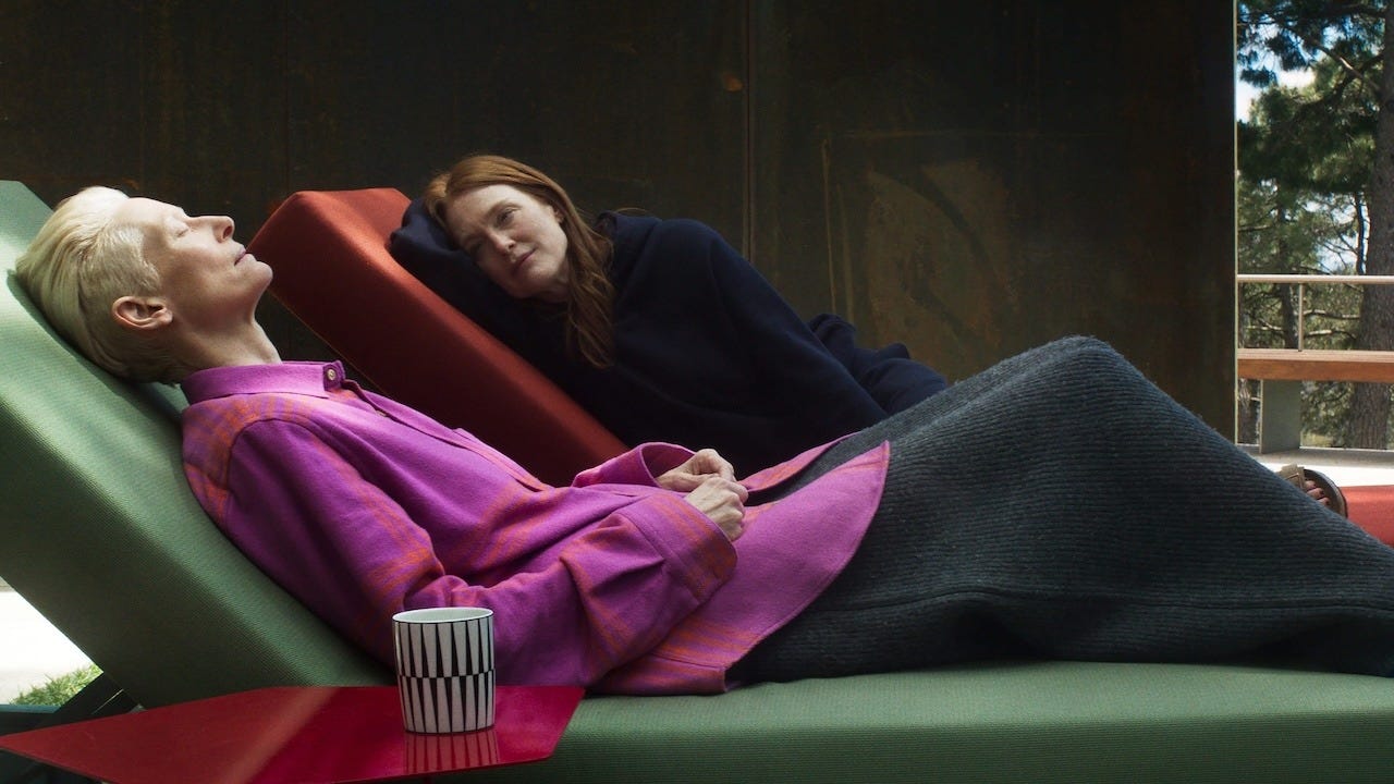 Still from the 2024 film The Room Next Door. Two women are shown in an intimate, contemplative moment. One is reclining on a green lounge chair wearing a vibrant pink blouse with orange accents and is partially covered by a dark gray blanket. The other woman, dressed in a navy blue outfit, rests her head on her hand while gazing at her companion. A striped black-and-white mug sits on a red side table in the foreground. The scene is set against a minimalist interior with large windows that offer a glimpse of trees outside.