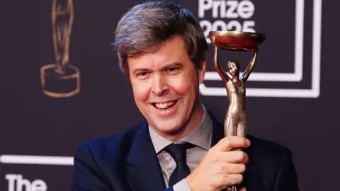 PA Media David Szalay smiling and holding up the Booker Prize trophy