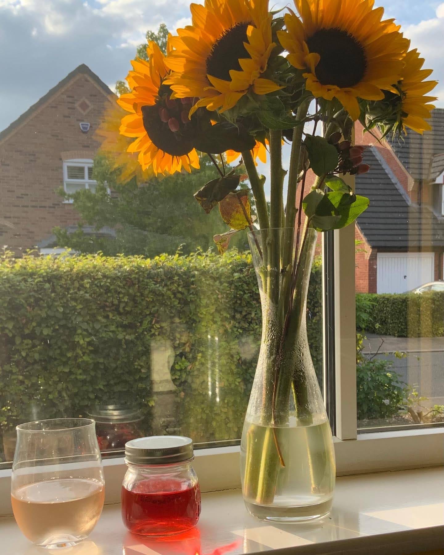 a glass of wine, a jar of rosepetal infused wine and a vase of sunflowers on a wondowsill a glass of wine, a jar of rosepetal infused wine and a vase of sunflowers on a wondowsill