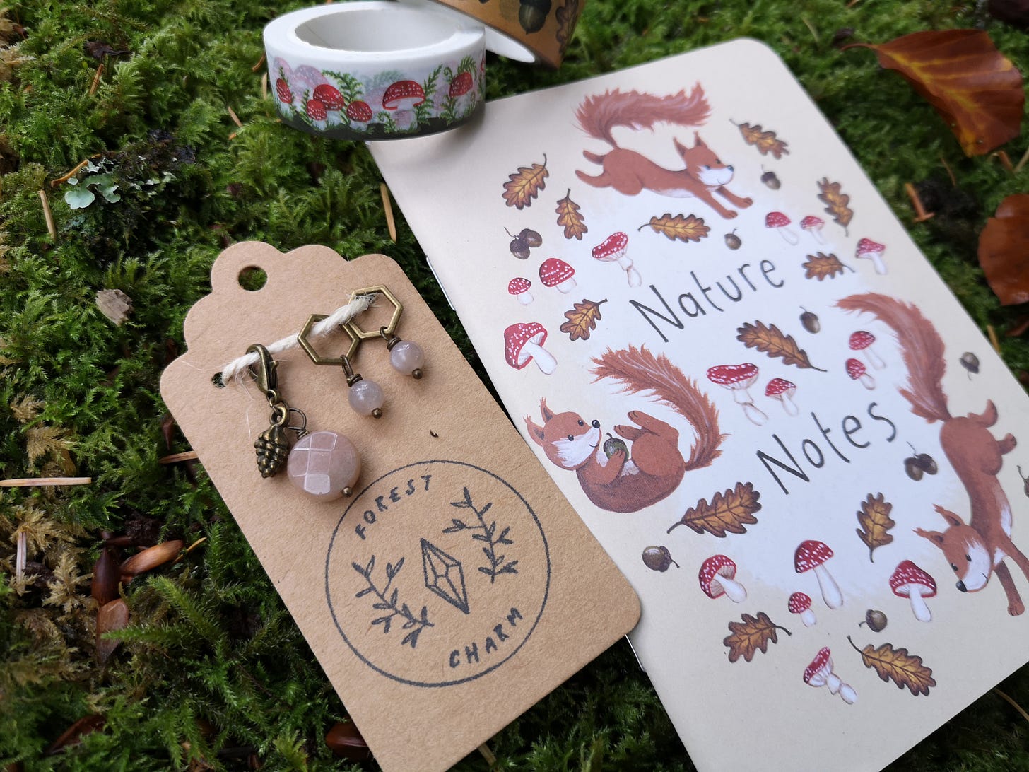 Image description: A bundle of goodies lying on some squishy green moss: a set of stitch markers and progress keepers featuring pink moonstone beads and a pine cone charm, lying next to a notebook with squirrels, mushrooms and oak leaves and some red and white mushroom washi tape.
