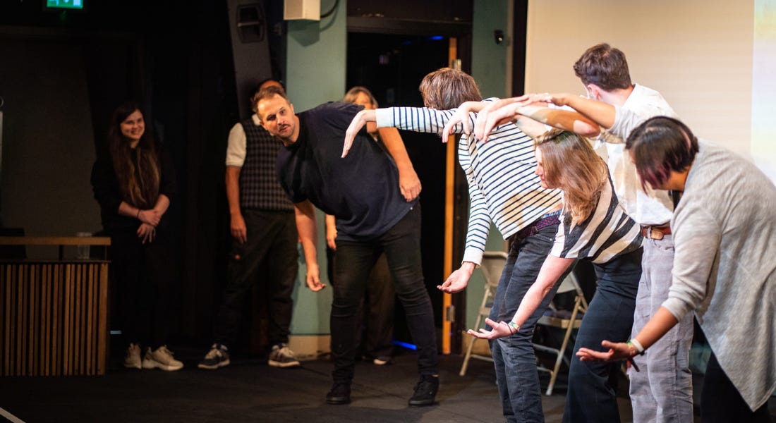 A group of people on stage with their left arms curved, looking to a man who is looking back A group of people on stage with their left arms curved, looking to a man who is looking back