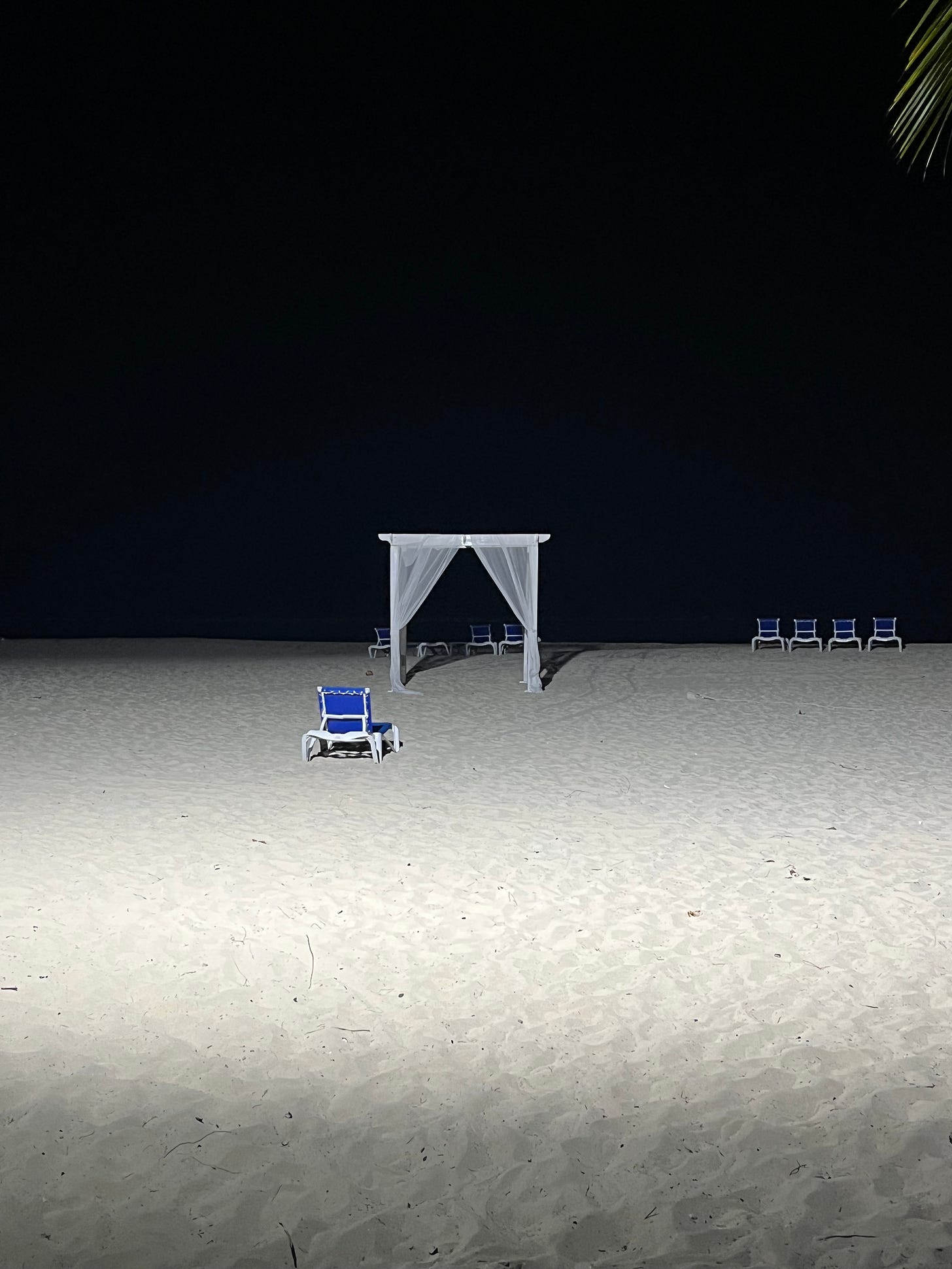 A white wedding arch on an abandoned beach with empty blue lounge chairs at nighttime.