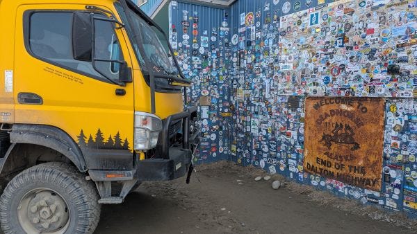 a big yellow truck  in front of a wall of stickers with a sign that reads Welcome to Deadhorse, Alaska, Top of the Dalton Highway