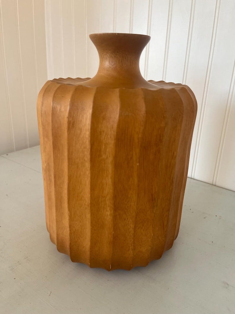 May include: A fluted, cylindrical wooden vase in a medium-brown tone. The vase has a slightly narrower neck and a wider body, with vertical grooves creating a ribbed texture.  The wood grain is visible, adding to the natural aesthetic. This mid-century modern style decorative vase is perfect for adding a touch of rustic elegance to any home decor.