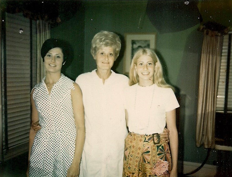 Vintage family photo of three women—mother, aunt, and grandmother—standing arm in arm in a mid-century living room, capturing a quiet, intimate moment across generations.