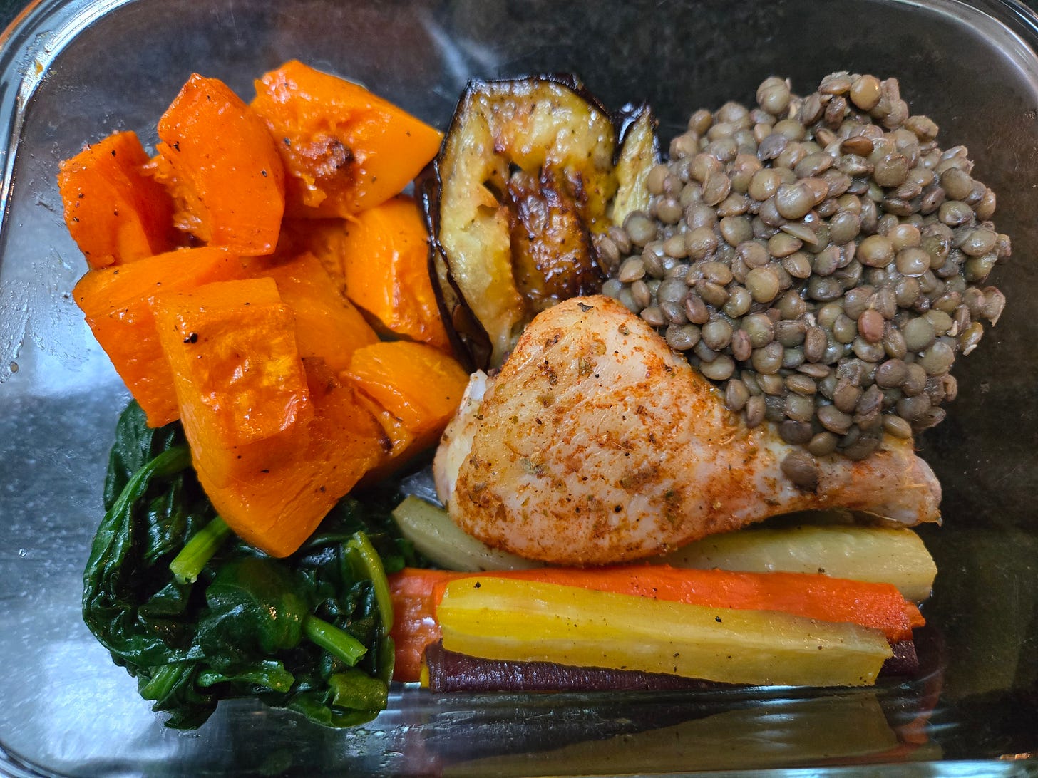 A rectangular glass container with roasted chicken, butternut squash cubes, carrots spears, and eggplant slices, with a pile of French lentils and sauteed spinach. 