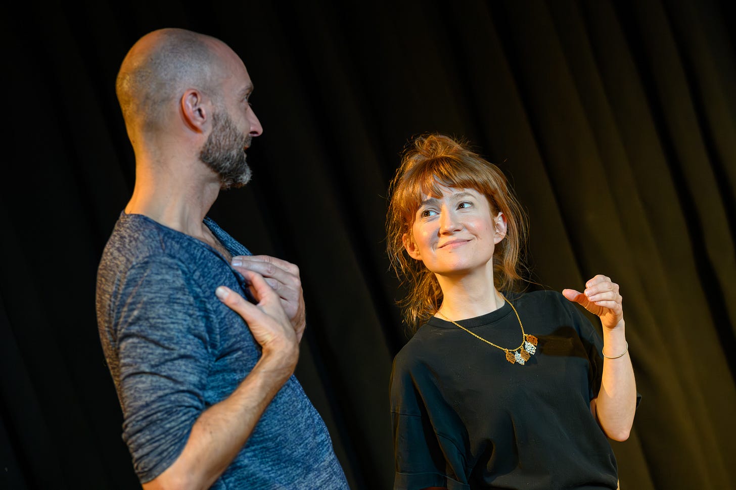 A man and woman stand next to each other on stage. The woman looks up at the man smiling. She is miming holding a glass