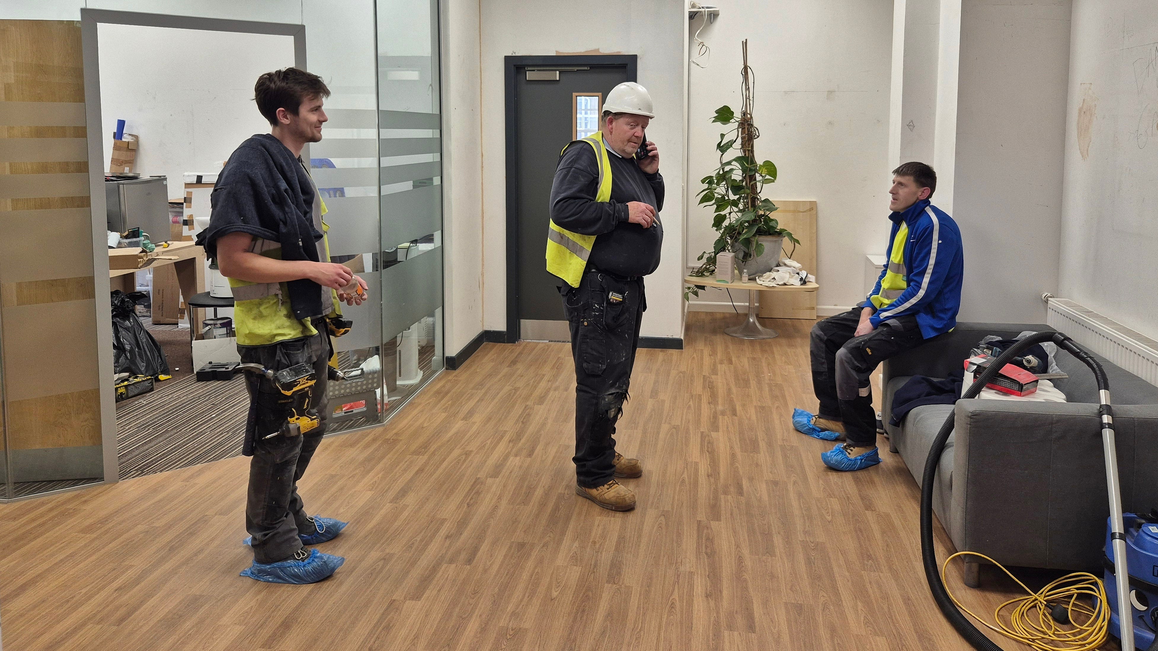 Three workmen, all with covers on their shoes and one in a hard hat, apply themselves to the outstanding construction isssues inside a white-walled meeting room at Bradford Arts Centre. The floor is of pale, Scand0-style wood and there is a glass-walled corner office full of moving-in clutter behind them.