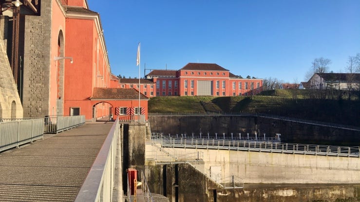 A 1930s hydro-electric dam built on the Rhine River. The buildings are painted a terracotta orange. A single-story structure, a former customs house, creates a gate over a wide walkway connecting the Swiss shore (depicted) with the German shore. The project was a Swiss and German collaboration.