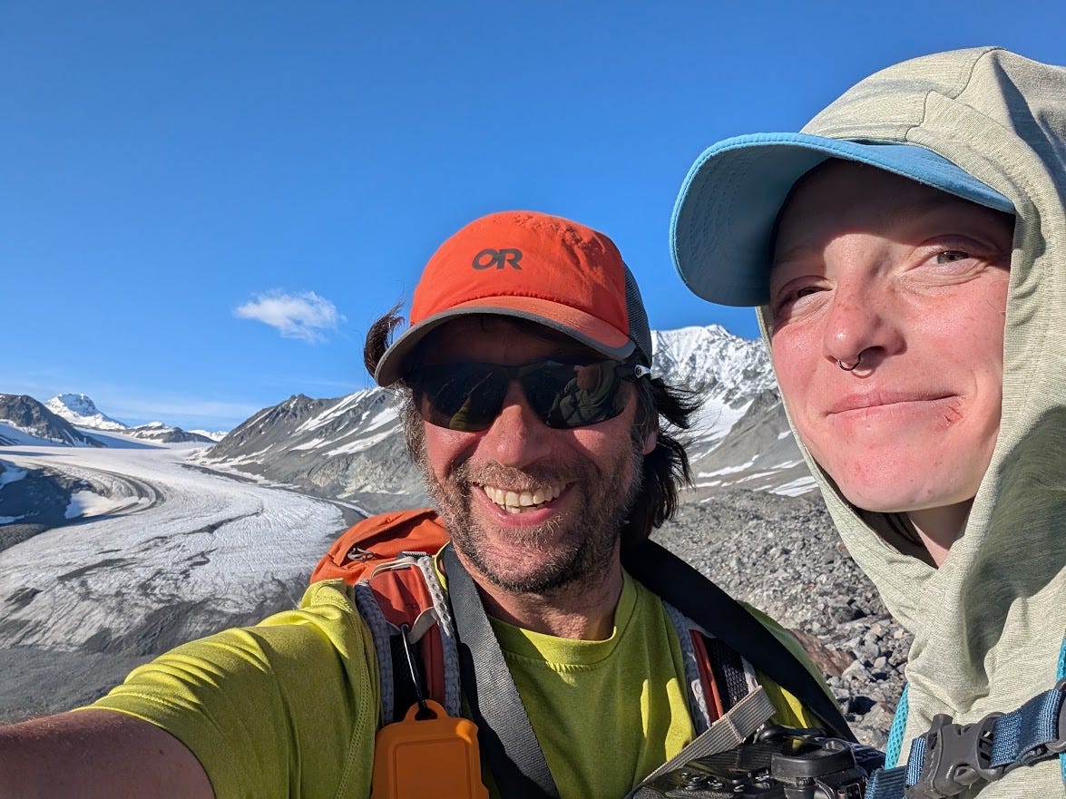 Two hikers pose close to the camera, one in an orange cap, sunglasses, and lime shirt, the other in a pale hood and cap, both smiling. Behind them the Gulkana Glacier sweeps down its valley between snow-streaked peaks under a bright blue sky.