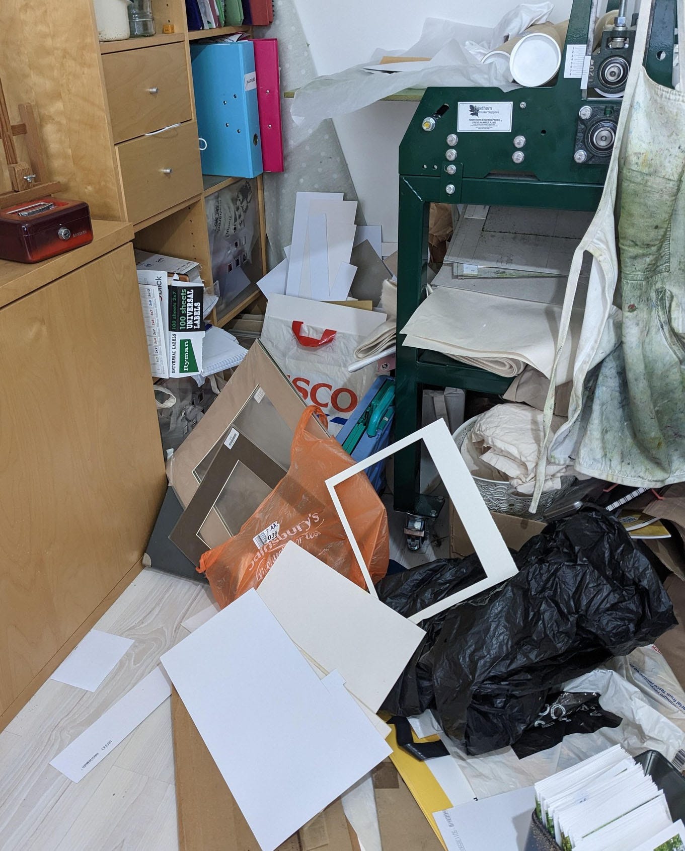 A corner of a room. The floorspace between a shelving unit and a printing press is covered in bags of junk.