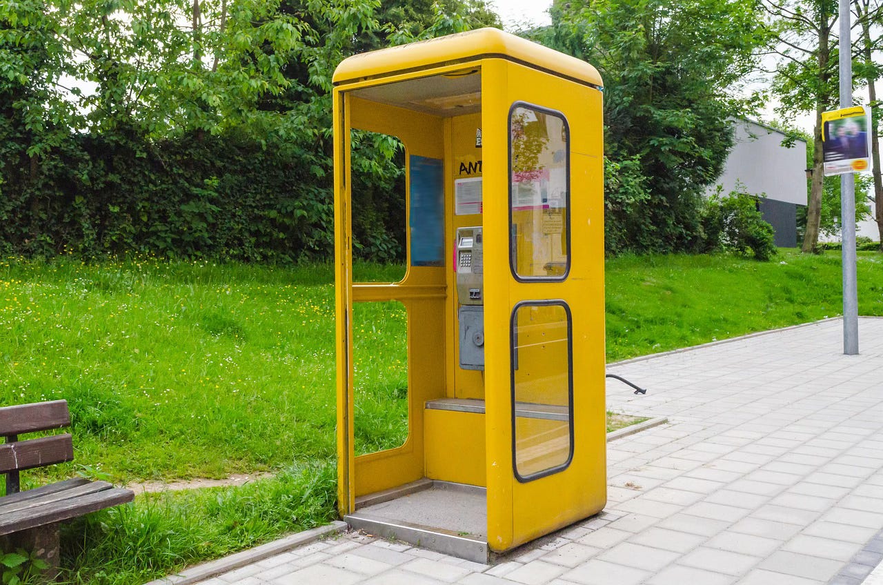 yellow public pay phone in a private booth with an inviting open door. Phone booth is located on a sideway, with a lush green lawn behind and an inviting park bench nearby.