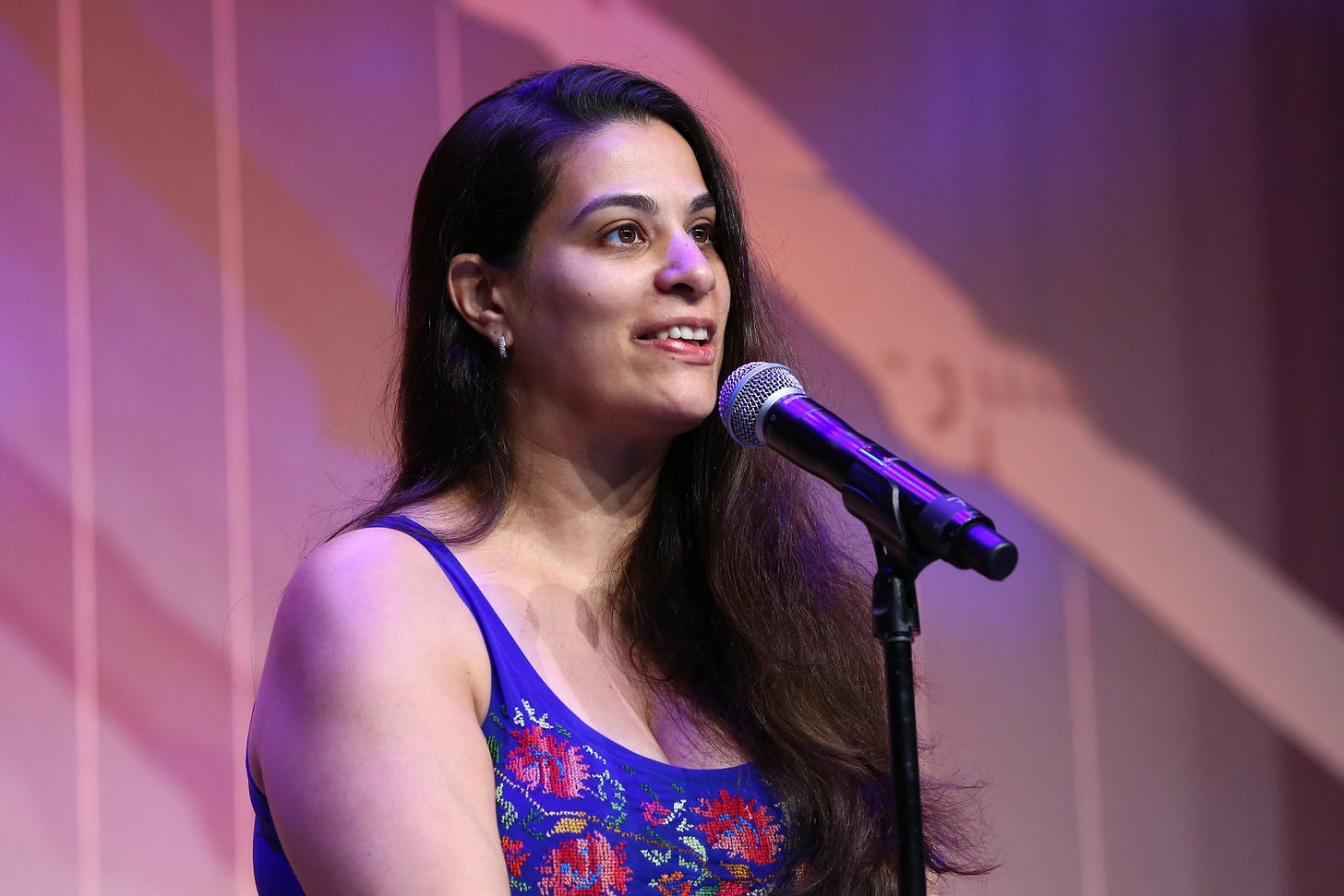 A photograph of Maysoon Zayid, an Arabic women with long black hair in a floral tank top in front of a microphone on some kind of a stage. 