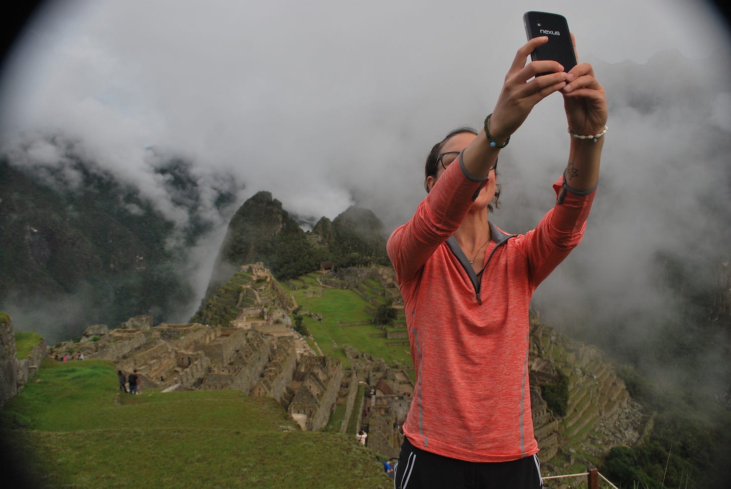 The picture shows the author, a white woman, standing at the top of the hill taking a selfie. She is wearing a pink shirt and posing in front of Machu Picchu in Peru.