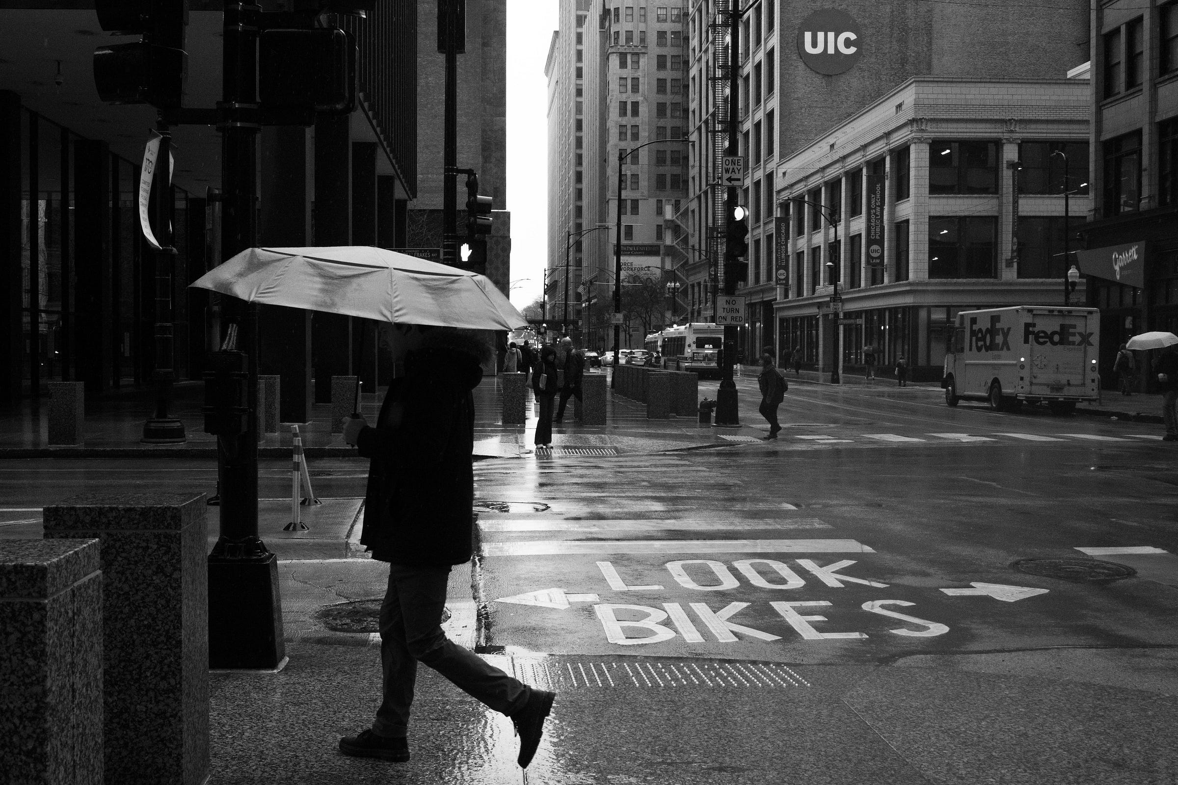 A person walks across a wet city street holding a large umbrella, with reflections on the pavement and a "LOOK BIKES" warning painted on the ground. Tall buildings, traffic, and pedestrians with umbrellas fill the background. A person walks across a wet city street holding a large umbrella, with reflections on the pavement and a "LOOK BIKES" warning painted on the ground. Tall buildings, traffic, and pedestrians with umbrellas fill the background.