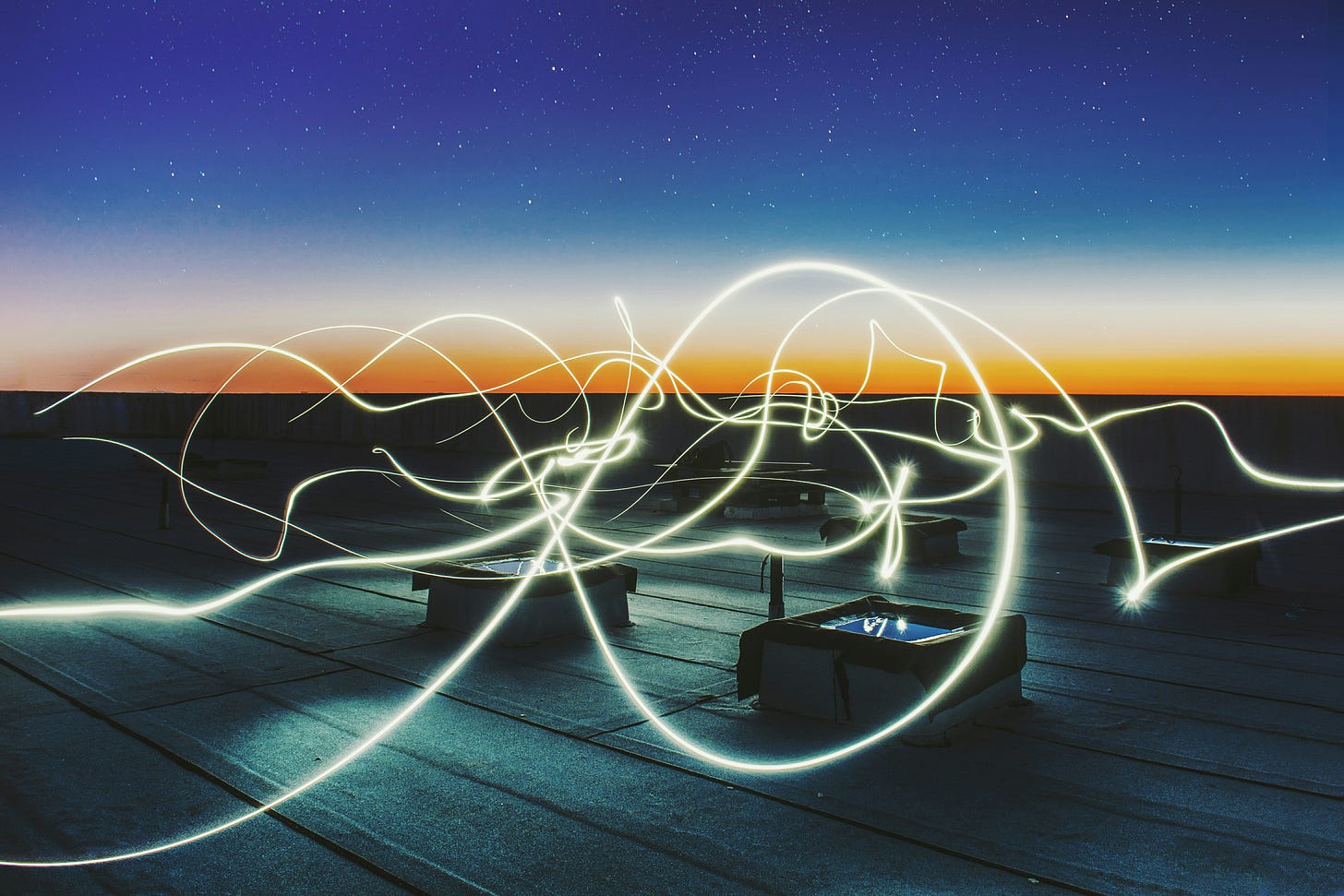 A rooftop scene at twilight with streaks of glowing light trails weaving through the air, creating a dynamic, energetic effect against a starry sky and sunset horizon. [Source: ChatGPT]