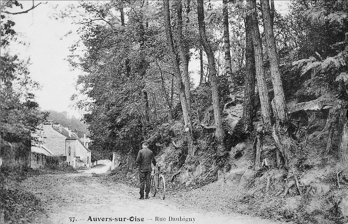 Postal com foto em preto e branco. No centro, aparece um homem de costas empurrando uma bicicleta. Ao seu lado direito, uma pequena encosta com árvores. Nela, as raízes são aparentes e bem similares às representadas por Van Gogh. Ao fundo da foto se vêem algumas casas.