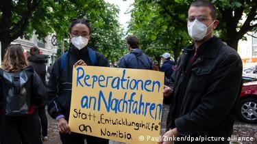 Two people holding a placard calling Germany to pay reparations to Namibia Two people holding a placard calling Germany to pay reparations to Namibia