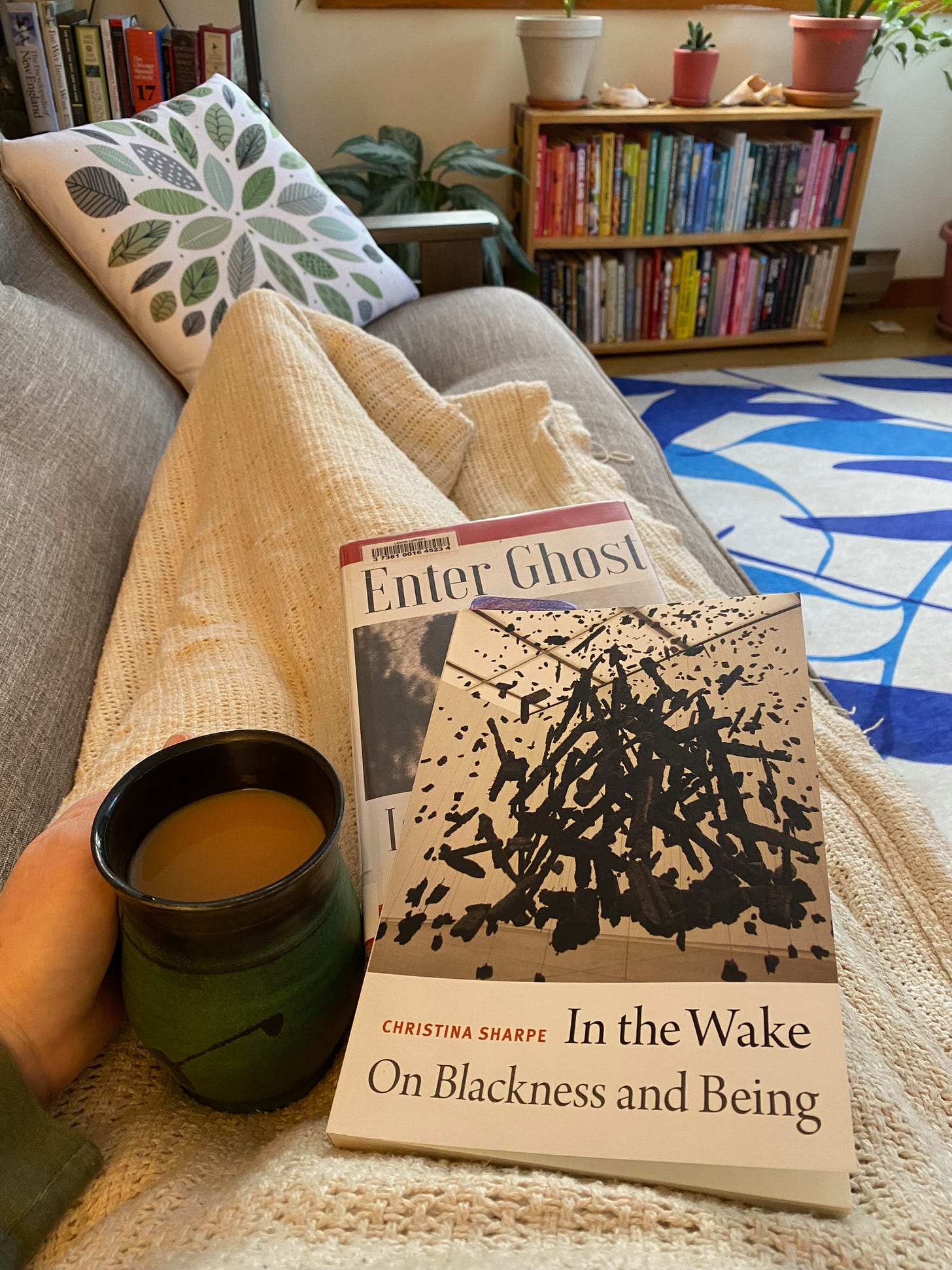 My legs stretched out under a blanket on a couch. I’m holding In the Wake, Enter Ghost, and a mug of tea on my lap.