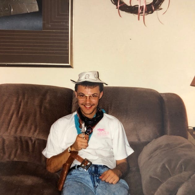 A vintage photo of a young man sitting on a brown plush couch, dressed in a playful cowboy outfit. He wears a white plastic cowboy hat, a white T-shirt with a pink gecko logo, a teal-and-black bandana around his neck, and a toy gun in a brown holster strapped over blue jeans. He smiles slightly while pointing the toy gun toward the camera. Behind him are a framed mirror and a wall wreath with pink ribbon.