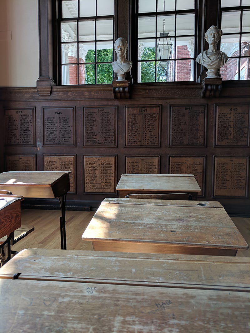 Photo of an old school room with wooden desks. There are names of graduates carved into the wall with their graduation year, and busts displayed above them. Photo of an old school room with wooden desks. There are names of graduates carved into the wall with their graduation year, and busts displayed above them.