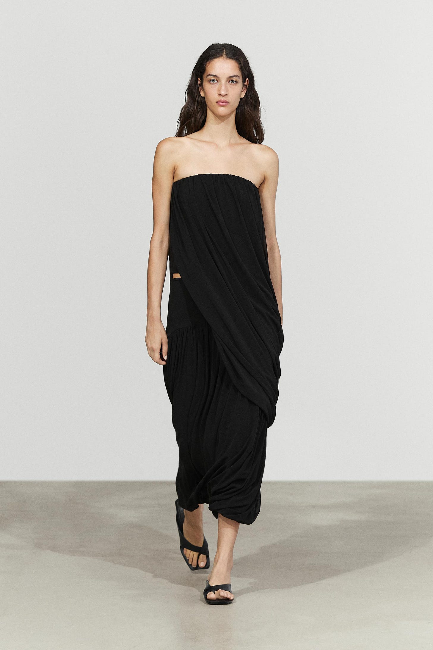 Ensemble composé d'un long haut noir à décolleté bustier, design drapé et ourlet asymétrique, d'une jupe midi noire drapée et de sandales noires.