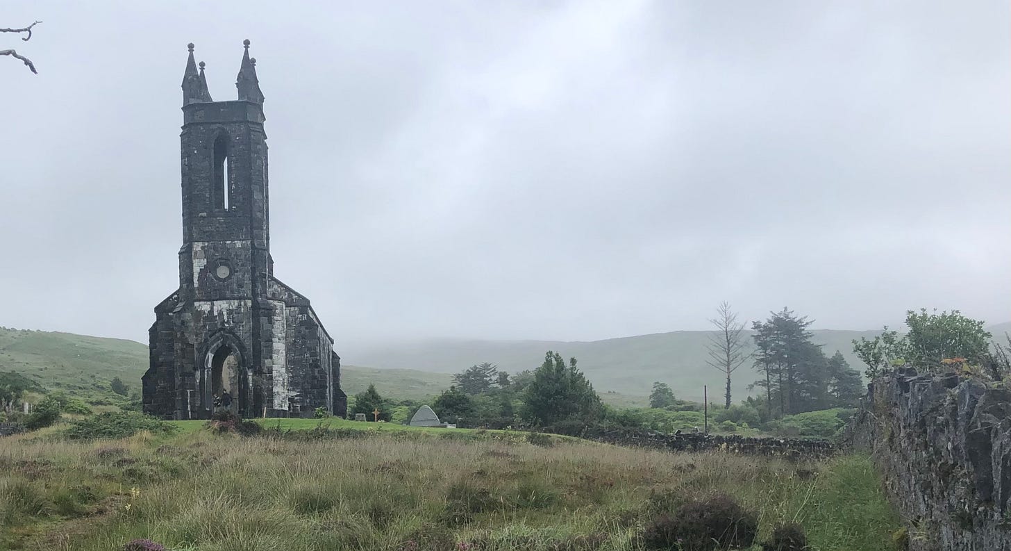 Dunlewey Church ruins in County Donegal, Ireland, viewed from across a misty field with surrounding hills and gravestones.