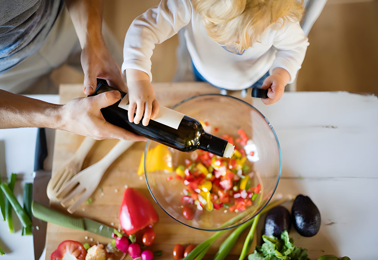 Parent and child cooking together, adding oil to a salad.