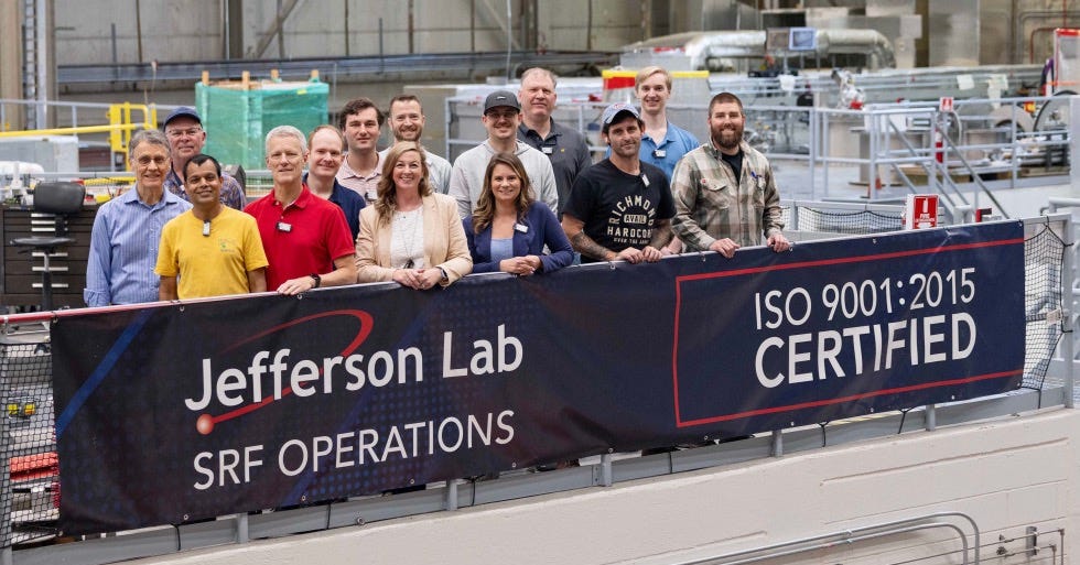 SRF Ops ISO 9001:2015 SRF Institute staff members stand behind a blue banner with "Jefferson Lab SRF Operations ISO9001:2015 Certified"