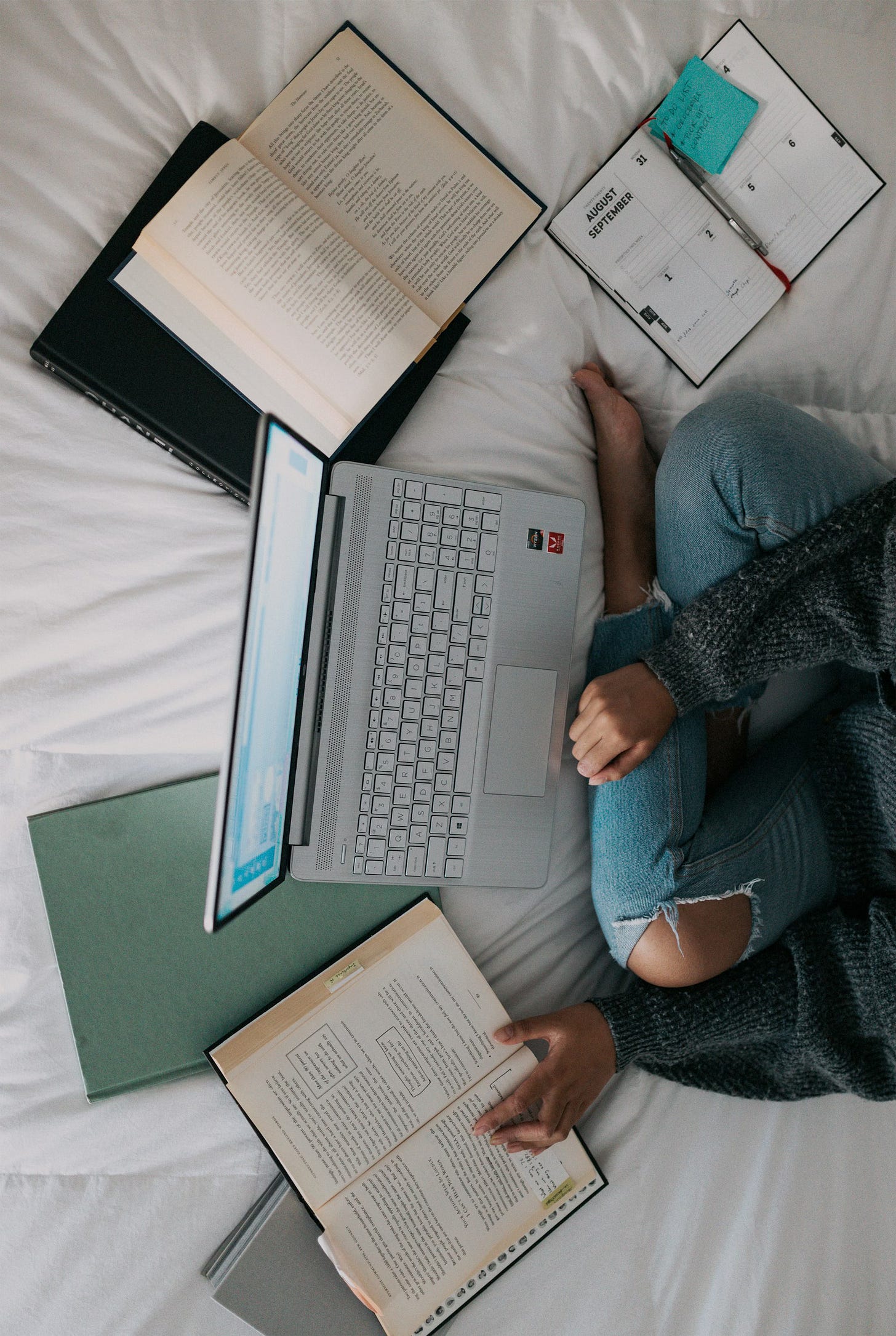 A girl sits cross-legged on a bed with a  white duvet to the right of the image, with a laptop and several books and a planner open in front of her. She's in jeans and a dark grey sweater.