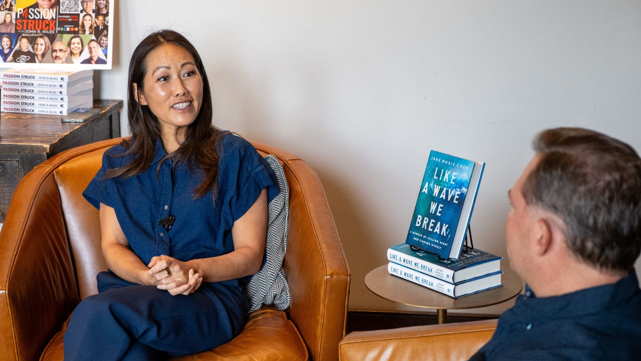 Jane Marie Chen, smiling softly and sitting in a leather armchair during a live Passion Struck podcast recording at the Oxford Exchange in Tampa. She wears a navy jumpsuit and has her hands gently clasped. On the small table beside her, a copy of her memoir ‘Like a Wave We Break’ stands upright, its wave-covered jacket clearly visible. In the background, a stack of ‘Passion Struck’ books and a podcast poster reinforce the intimate, warm setting.