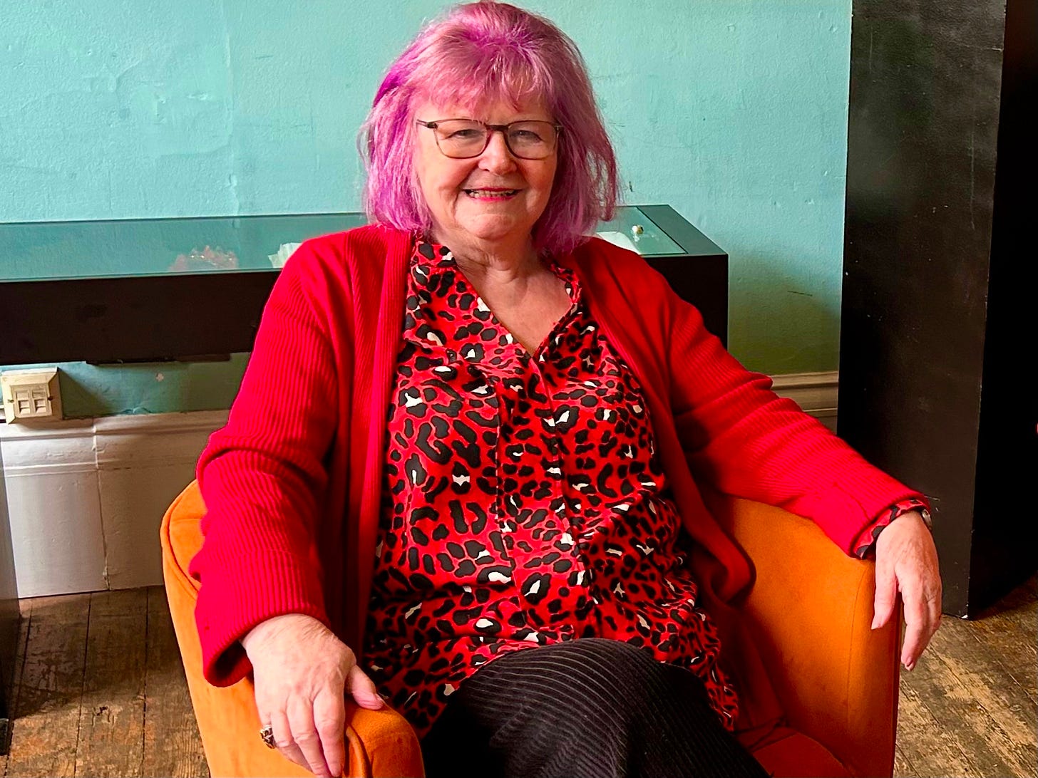 A vibrant portrait of Jeanne, a woman with bright purple hair and glasses, wearing a red leopard-print blouse and a red cardigan. She is smiling warmly while sitting in a tan leather armchair against a pale green wall.