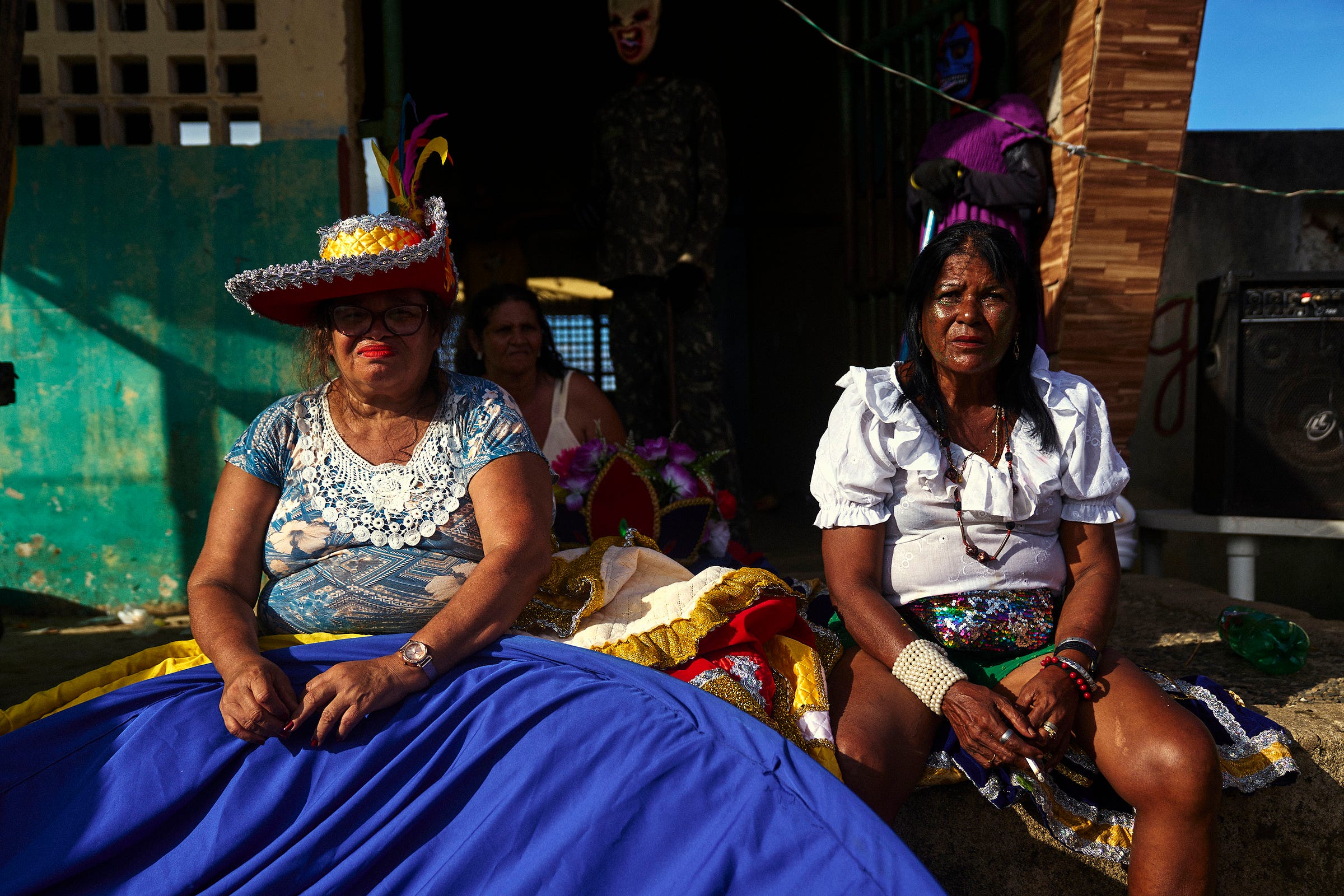 Two women in colorful costumes sit outdoors in bright sunlight. One wears a large red hat with feathers and a blue skirt; the other, in a white blouse and skirt, has a beaded bag. Others and a masked figure are in the shaded background.