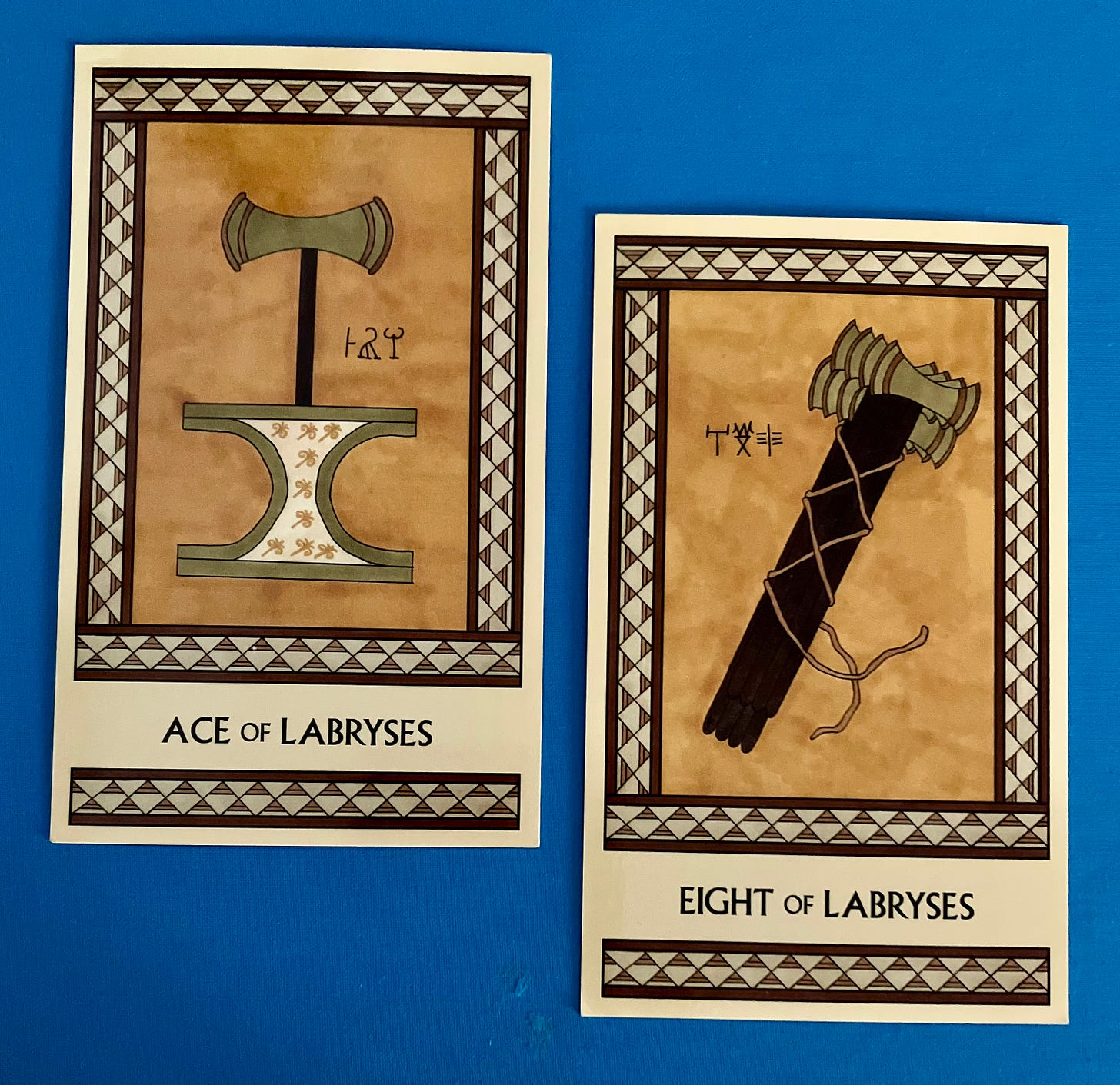 Two Minoan Tarot cards on a sky blue background. Both cards are in shades of gold and tan. The Ace of Labryses shows a labrys standing atop an incurved altar decorated with lilies. The Eight of Labryses shows a bundle of eight labryses that appear to be tied tightly together, until you notice that the ropes aren't actually knotted but have free ends. 