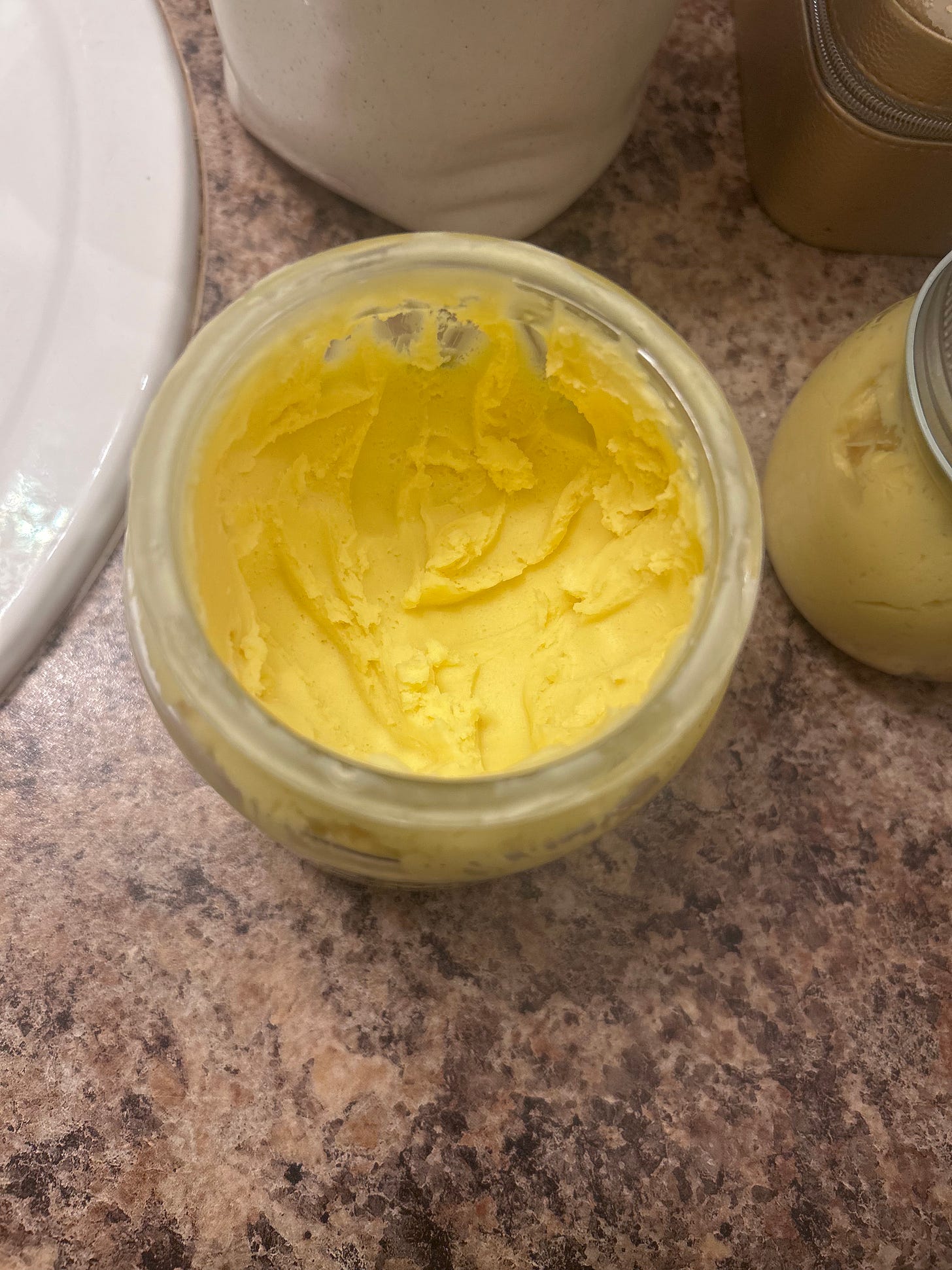 A close-up, top-down view of an open glass jar revealing the rich, whipped texture of DIY yellow shea body butter. The consistency is thick, creamy, and soft, showing the successful results of using arrowroot powder to create a non-greasy finish.