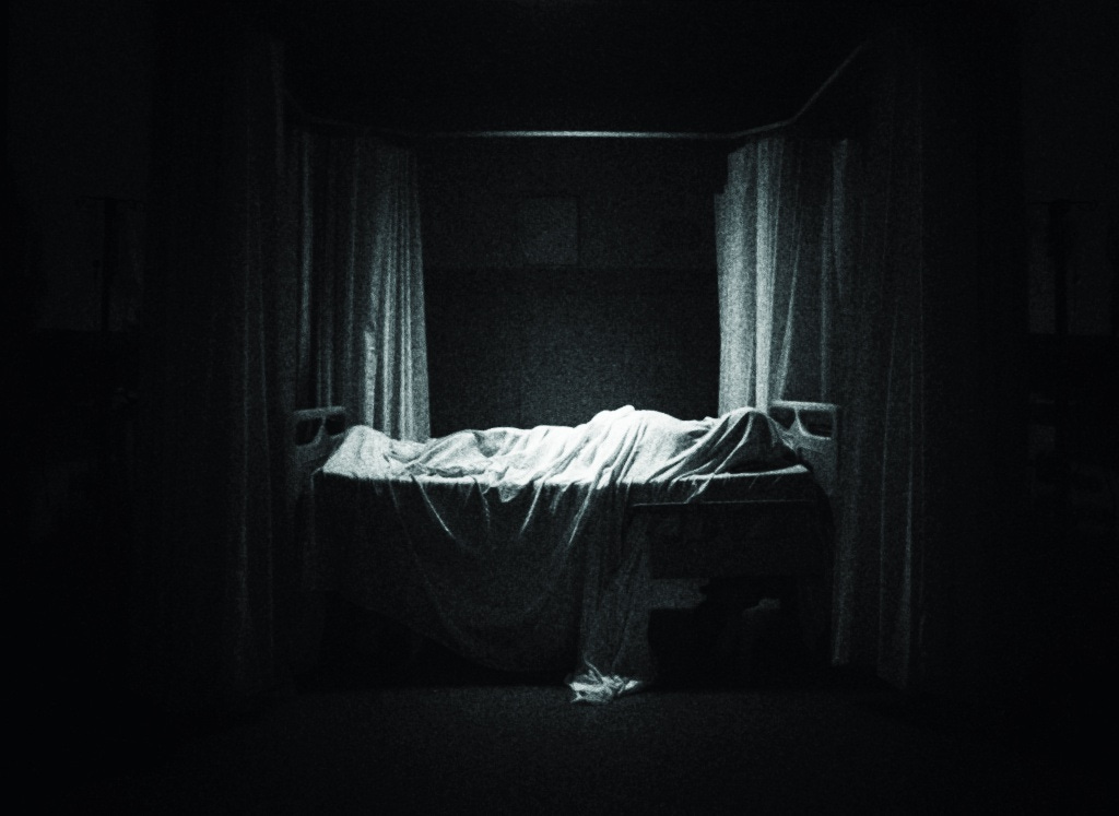 A figure sleeps underneath a sheet in a hospital bed in a dark room