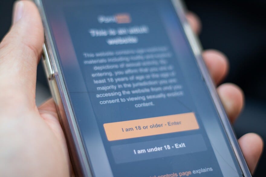 A hand holds a smartphone with age verification text on a pornographic website. 