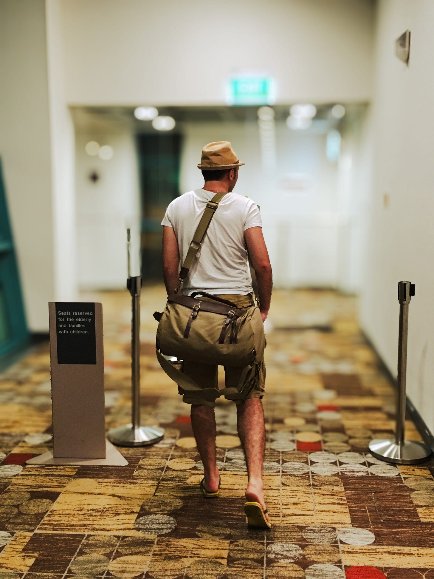 Un uomo di spalle, con uno zainetto, si avvia verso il check-in nel corridoio di un aeroporto. Indossa sandali, bermuda, una maglietta bianca e un cappello.