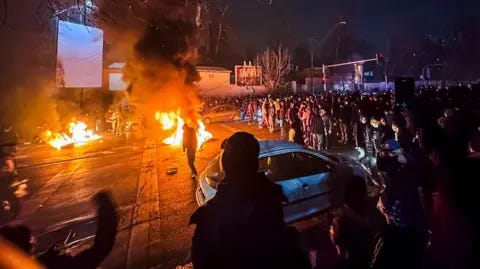 MAHSA / Middle East Images / AFP via Getty Images Iranians gather while blocking a street during a protest in Tehran, Iran on January 9, 2026. Masked people stand on the road as they watch two fires burn on the pavement in the dark. MAHSA / Middle East Images / AFP via Getty Images Iranians gather while blocking a street during a protest in Tehran, Iran on January 9, 2026. Masked people stand on the road as they watch two fires burn on the pavement in the dark.