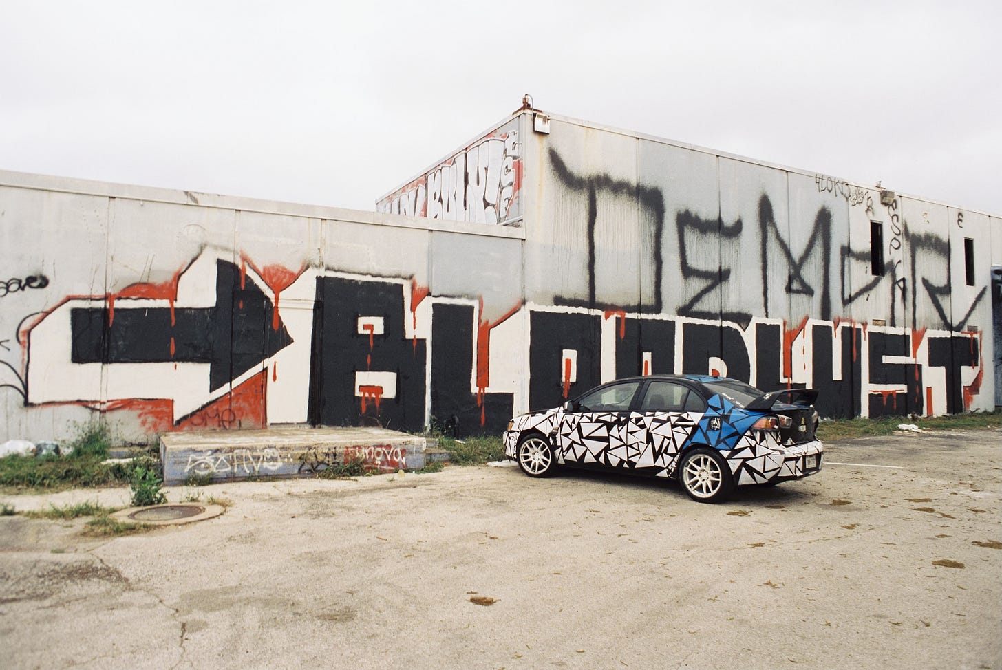 Late model sport sedan covered in triangle decals parked in front of abandoned factory with giant graffiti that reads ">BLOODLUST"