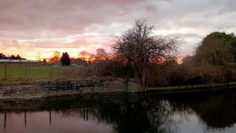 Three images, the first is just the end of a pink narrow boat in front of the canal water, with a canal path also in view. Second image is of the top of a narrow boat with solar panels, it is inside a lock, with the lock edges clearly visible including brick walls and trees above. Thirdly there is a picture of a sunset over the canal with the reflection in the still water.