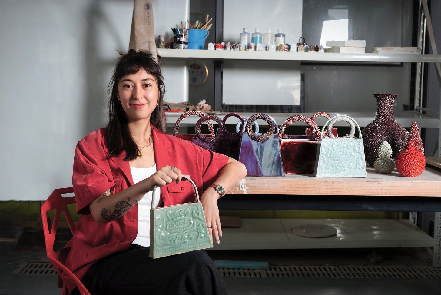 Anne Loquineau sits in her studio holding one of her porcelain handbags, with a display of her colorful ceramic works arranged on the table behind her.