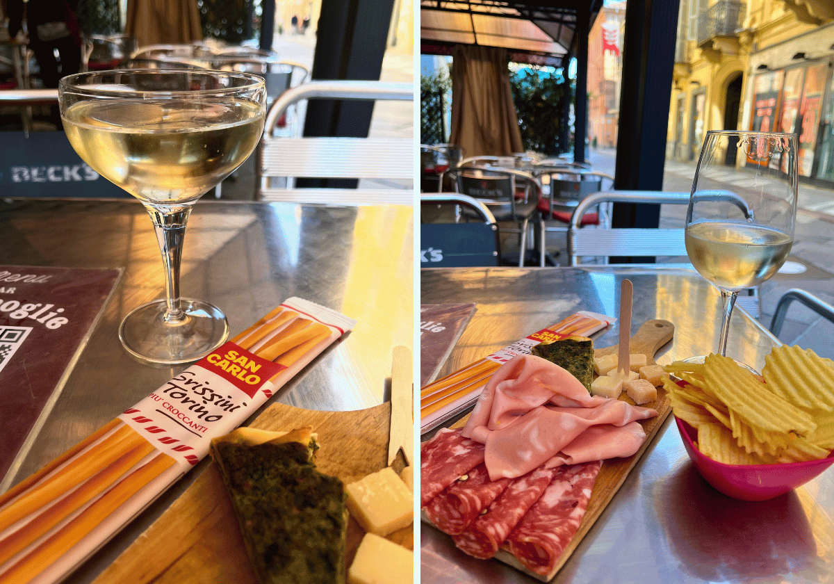 Two glasses of white wine on a metal table with packaged breadsticks, a slice of savory tart, mortadella, and potato chips. Two glasses of white wine on a metal table with packaged breadsticks, a slice of savory tart, mortadella, and potato chips.