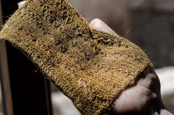 A hand holds a delicate piece of lace knitted textile made from the silk of a mussel — known as sea silk — in its natural olive green colour.