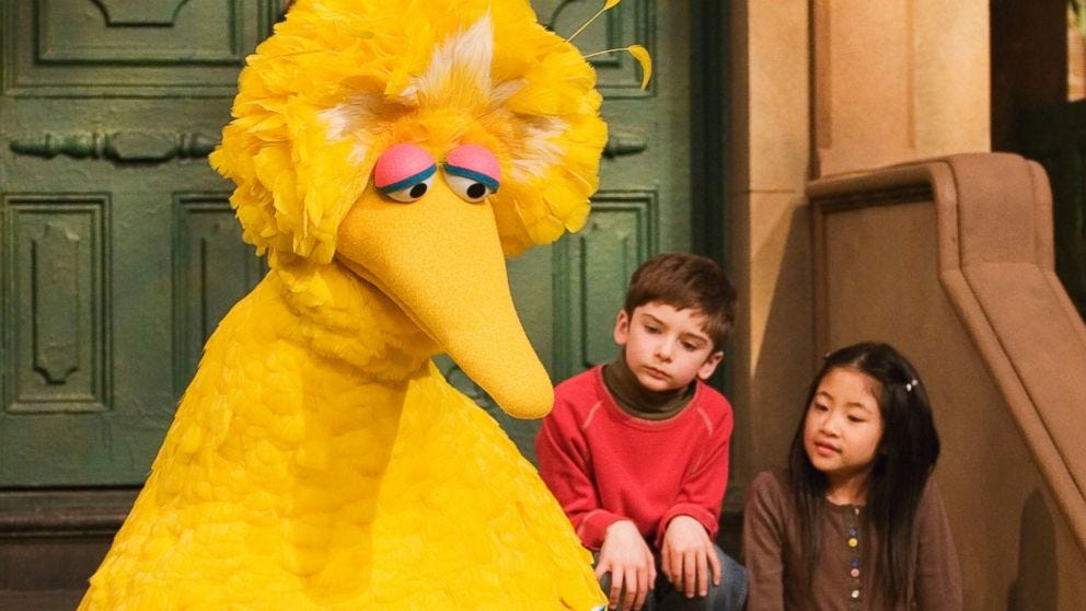 Big Bird sits on a stoop on Sesame Street next to two small children. He looks sad.