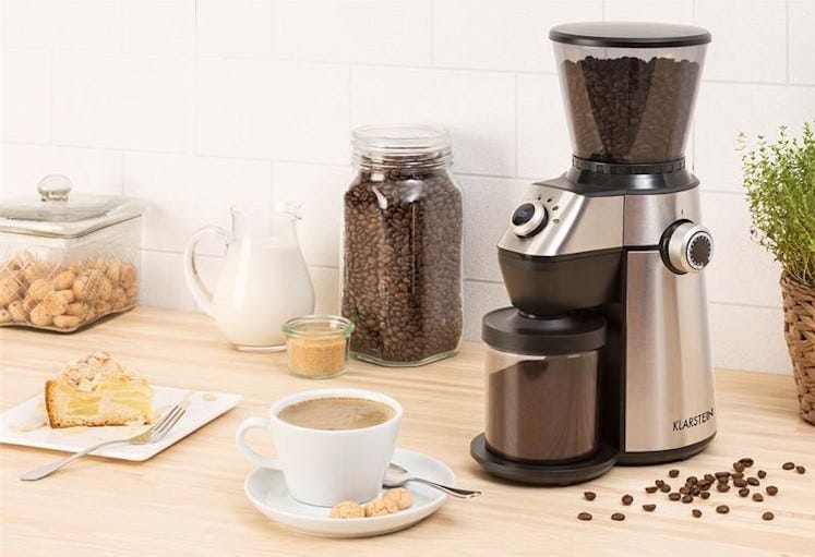 El molinillo en cuestión sobre encimera de madera, junto a una taza de cafá con leche, varios dulces, jarra de leche...